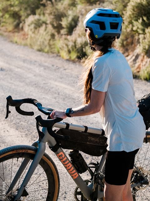 Eine Person mit blauem Helm und weißem Hemd steht neben einem grauen Fahrrad auf einem sonnenbeschienenen Schotterweg mit Grün im Hintergrund.