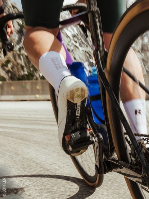 Nahaufnahme des Fußes eines Radfahrers auf einem Pedal, mit weißem Radschuh, blauer Wasserflasche und verschwommener Bewegung auf einer Straße.