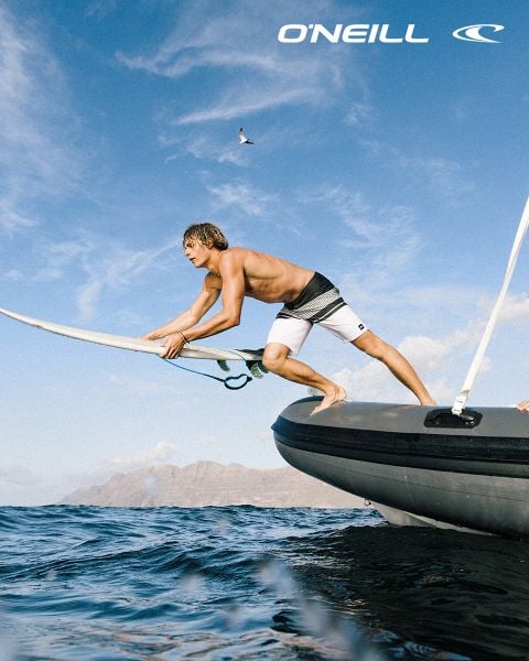 Ein Mann in Badehose hält ein Surfbrett auf einem Schlauchboot und bereitet sich auf das Surfen vor, mit dem Meer und den Bergen im Hintergrund; darüber das O'Neill-Logo.