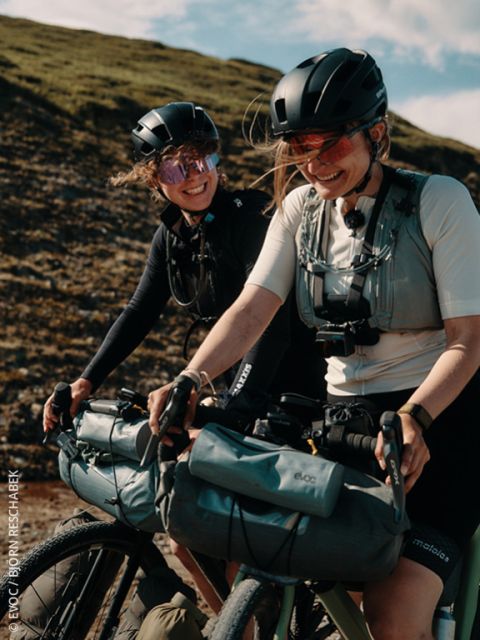 Zwei Frauen mit Helmen und Sonnenbrillen fahren lächelnd auf Fahrrädern im Freien, an denen Taschen befestigt sind. Im Hintergrund ist ein grasbewachsener Hügel zu sehen.