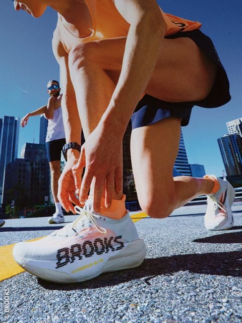Ein Läufer kniet auf einer Straße in der Stadt nieder, um seinen weißen Brooks-Laufschuh zu schnüren; im Hintergrund sind ein weiterer Läufer und hohe Gebäude zu sehen.