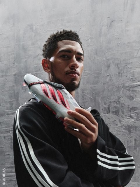 Ein Mann in einem Adidas-Trainingsanzug hält einen grau-roten Adidas-Fußballschuh vor sein Gesicht und steht vor einem strukturierten grauen Hintergrund.