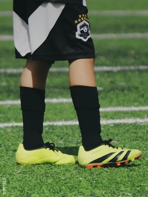 Ein kleiner Junge kickt auf einem Feld einen blauen Fußball, während ein Mädchen in Schuluniform im Hintergrund zusieht.