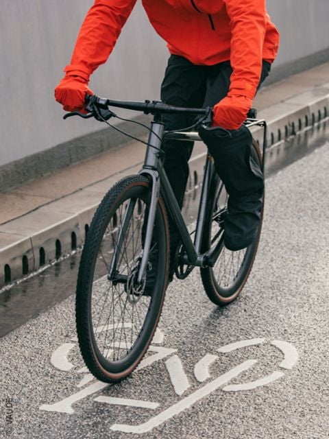 Ein Mann, der einen Helm, eine Sonnenbrille und Geschäftskleidung trägt, fährt mit einem Fahrrad mit Gepäcktaschen durch eine Straße in der Stadt, wobei der unscharfe Hintergrund die Bewegung anzeigt.