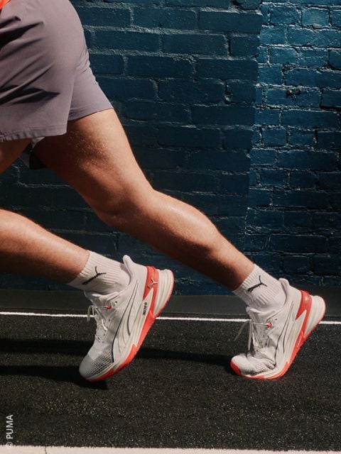 Eine Frau in einem blau-weißen Trainingsanzug sitzt auf dem Boden und posiert mit einem angehobenen Knie. Sie trägt weiße Socken und silberne Under Armour Turnschuhe und lächelt leicht vor einem schlichten hellen Hintergrund.