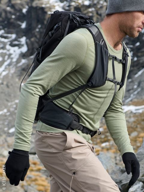 Ein Mann in einem grünen Hemd, einer beigen Hose und Handschuhen, der in felsigem, bergigem Gelände wandert und einen Rucksack sowie eine graue Mütze trägt.