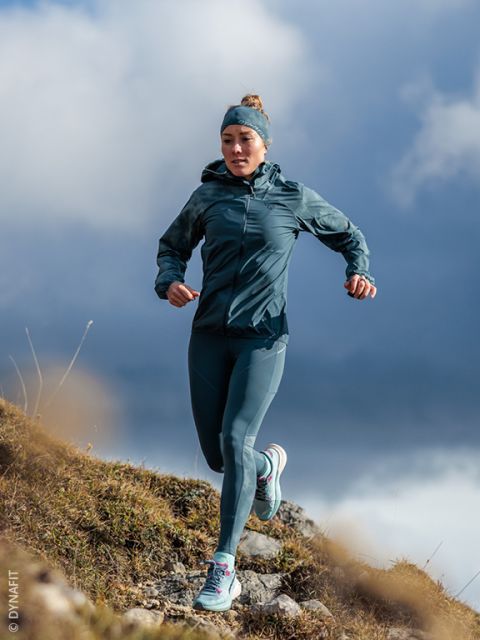 Eine Frau in türkisfarbener Sportbekleidung läuft auf einem felsigen Pfad bergab, im Hintergrund ist ein bewölkter Himmel zu sehen.