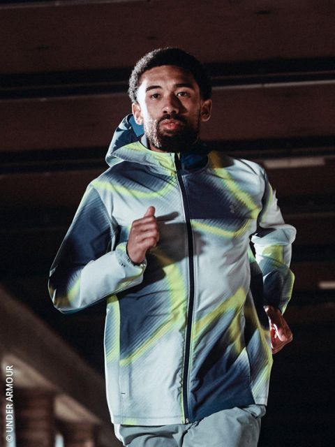 Eine Person, die eine Kapuzenjacke und Laufschuhe trägt, joggt an einem kalten, klaren Tag einen verschneiten Weg entlang, mit der Skyline der Stadt im Hintergrund.