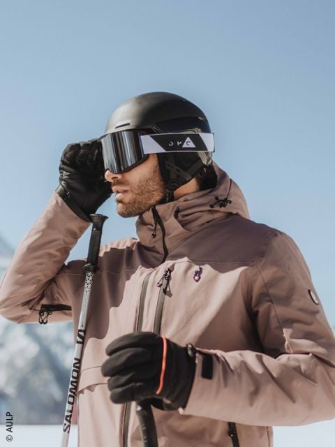 Nahaufnahme einer Person, die einen grauen Skihelm, eine reflektierende Skibrille und eine dunkle Jacke trägt, mit einer verschwommenen Schneelandschaft im Hintergrund.