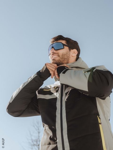 Ein Skifahrer, der einen weißen Helm, eine reflektierende Brille, eine schwarz-braune Jacke und Handschuhe trägt, schließt seine Jacke im Freien mit verschneiten Bergen im Hintergrund ab.